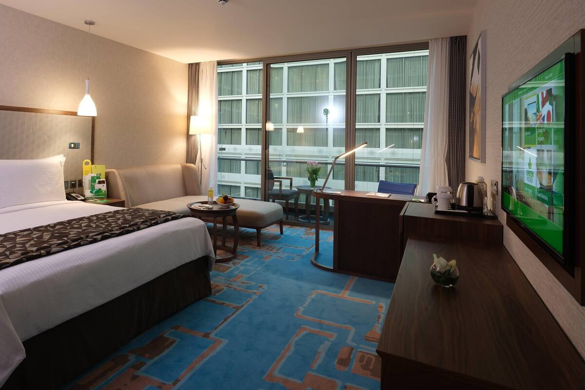 A hotel room with a large bed, a desk, a chair, a small table with a lamp, and a TV on the wall.