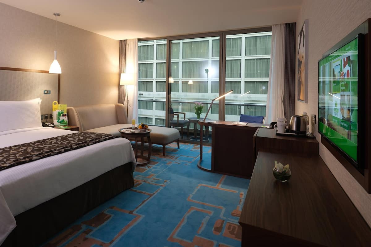A hotel room with a large bed, a desk, a chair, a small table with a lamp, and a TV on the wall.