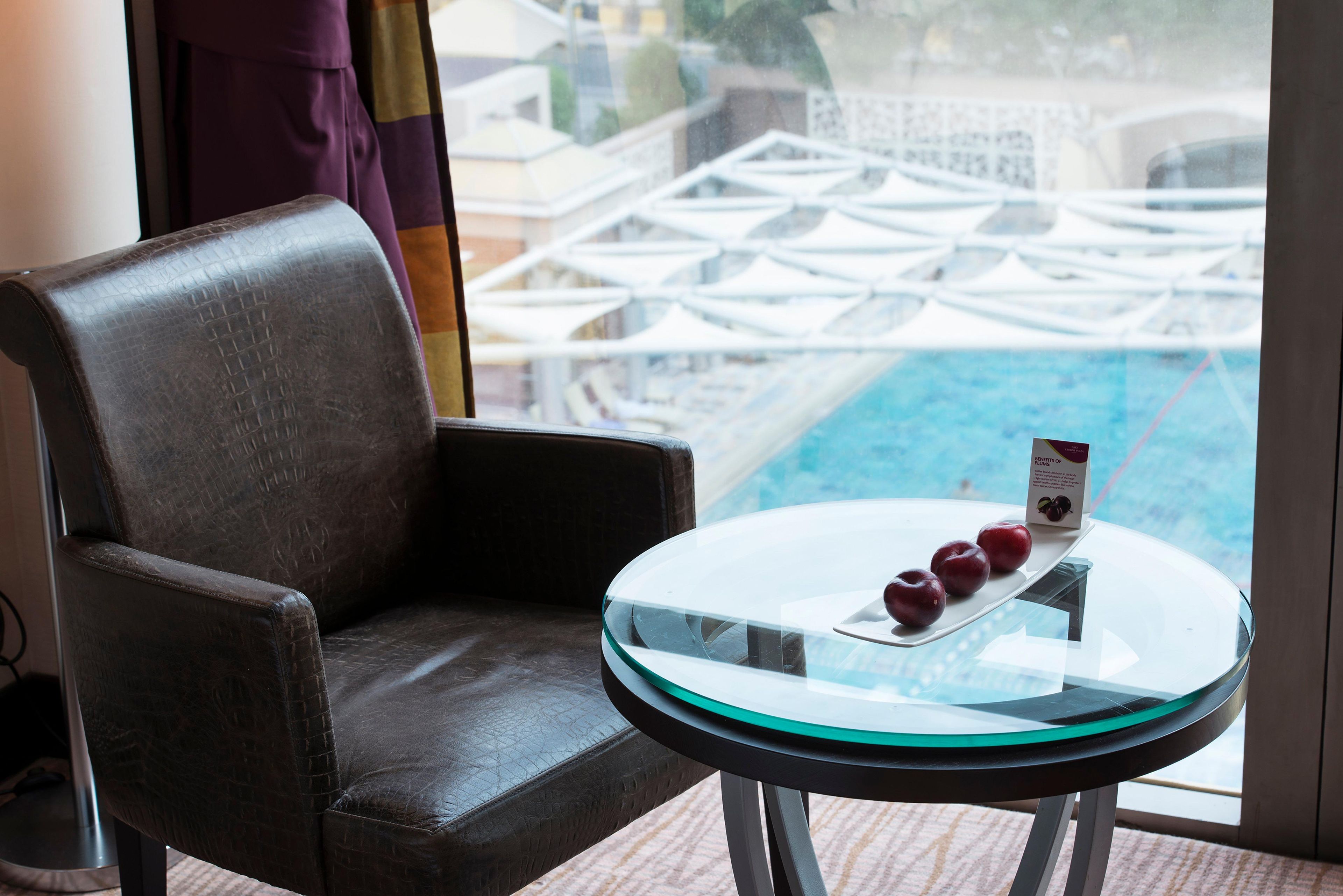 Un sillón de cuero y una mesa de vidrio con una bandeja de fruta en una habitación con vista a una piscina y sombrillas.