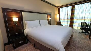 A hotel room with a large bed, two bedside tables with lamps, a chair, and a view of the ocean through large windows.