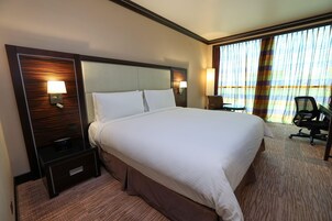 A hotel room with a large bed, two bedside tables with lamps, a chair, and a view of the ocean through large windows.