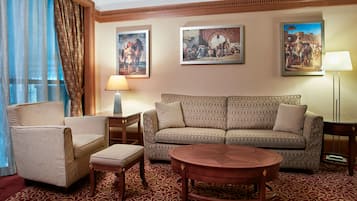 A hotel room with a sofa, armchair, ottoman, and a round coffee table. There are two paintings on the wall and a lamp on the side table.