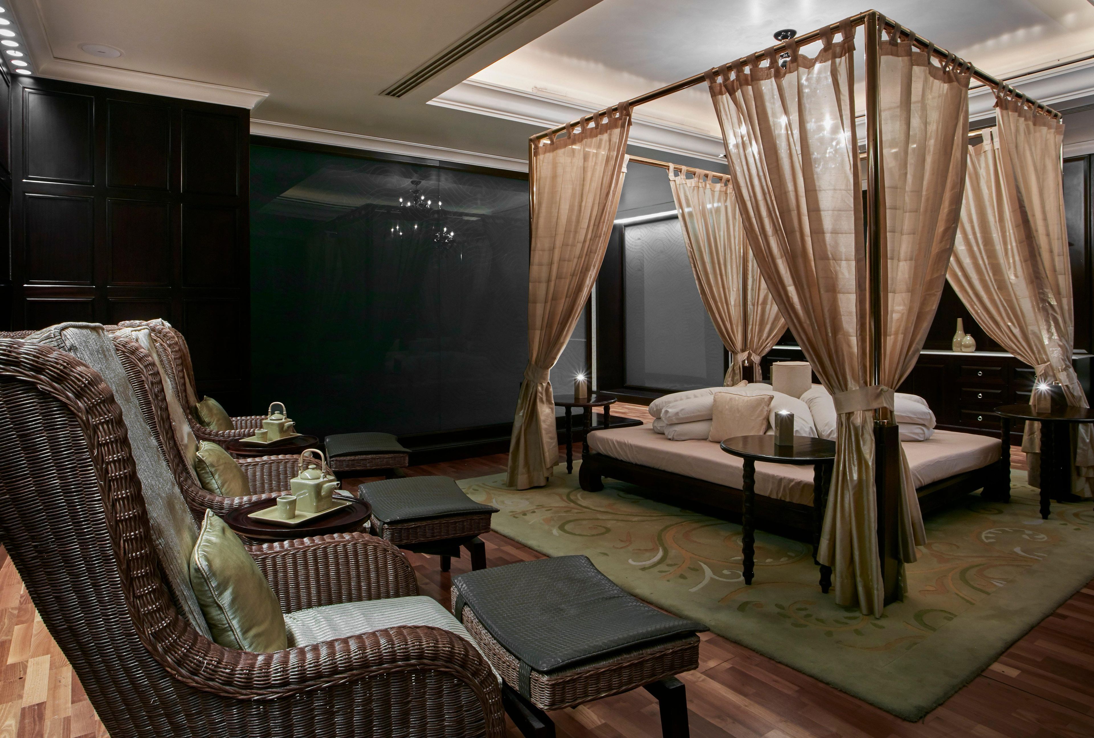 A room with a four-poster bed, a wicker sofa, a small table with a tray, and a green patterned rug.