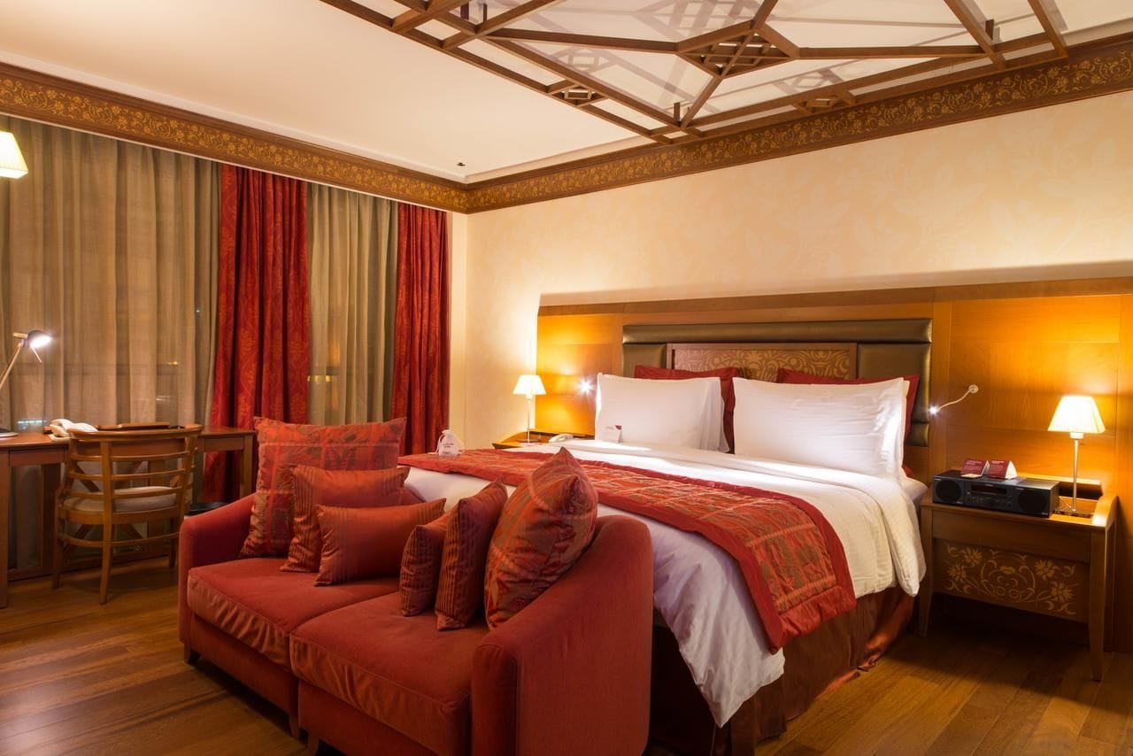 A hotel room with a large bed, a sofa, a desk with a lamp, and a nightstand with a lamp.