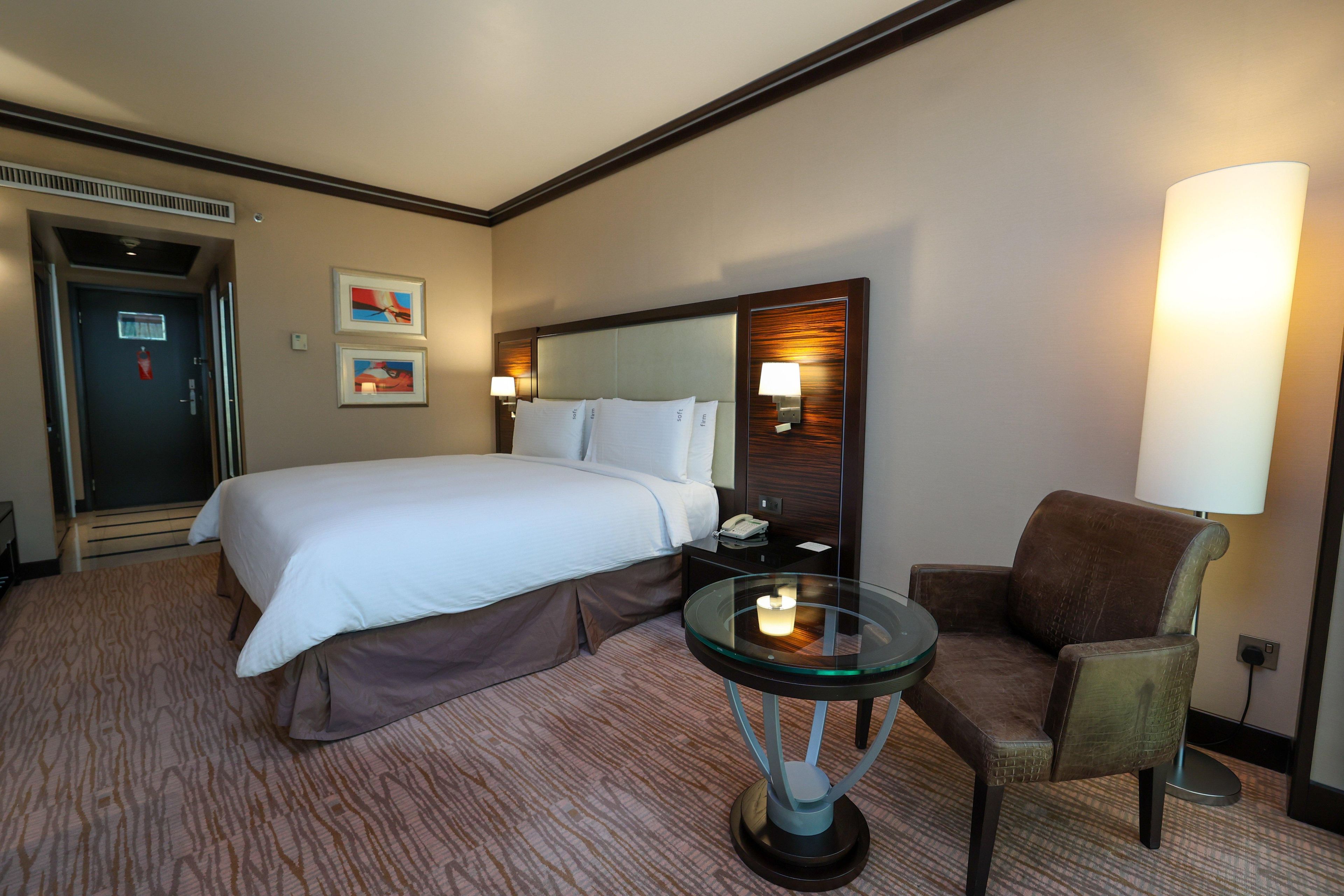 A hotel room with a bed, a chair, a glass table with a candle, a lamp, and artwork on the wall.