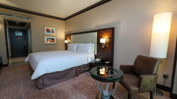 A hotel room with a bed, a chair, a glass table with a candle, a lamp, and artwork on the wall.
