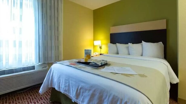 A hotel room with a bed, a bedside table, a lamp, and a window with curtains. - Dulles Suites (Sterling)