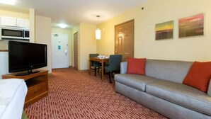A hotel room with a television, a sofa, a dining table with chairs, a microwave, and a patterned carpet. - Dulles Suites (Sterling)