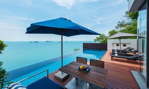 Una piscina infinita con vistas al mar, una terraza de madera con una mesa y sillas, y un área de descanso con sombrillas.
