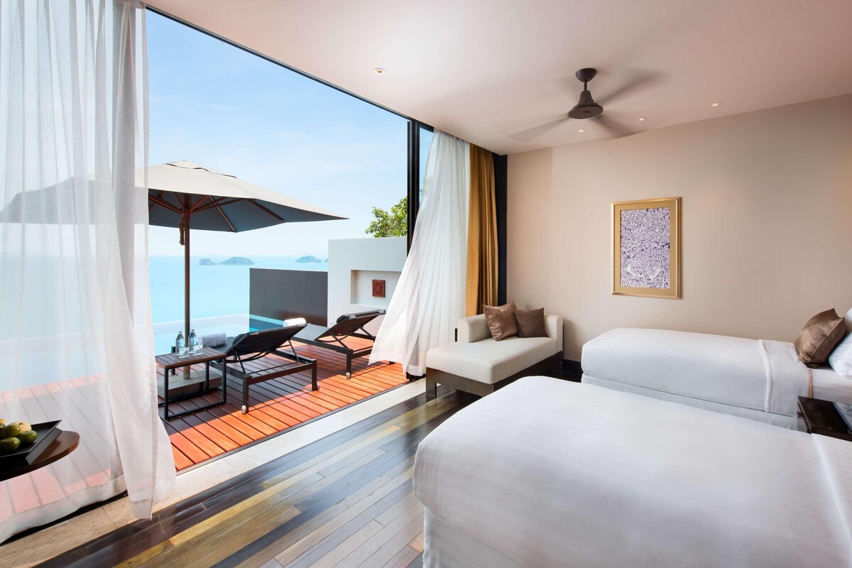 A hotel room with a large window, a ceiling fan, a bed, a sofa, and a small table with a lamp.
