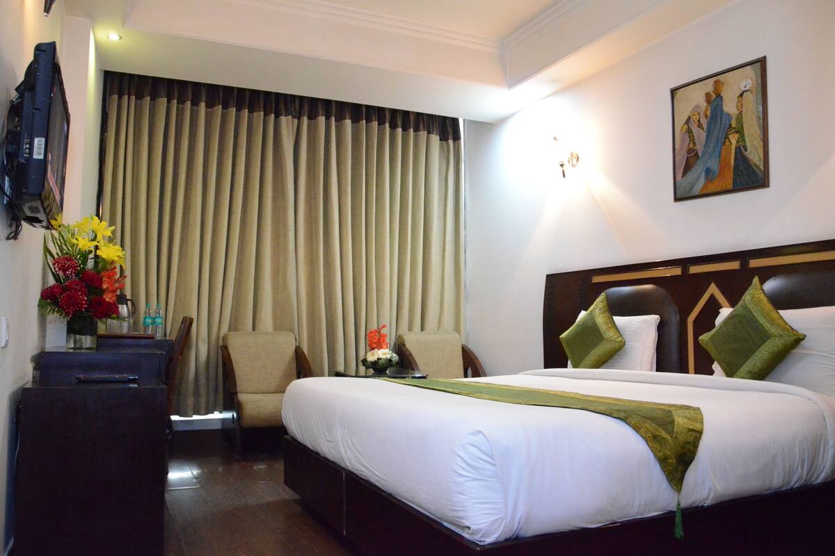 A hotel room with a bed, a chair, a desk, a TV, and a painting on the wall.