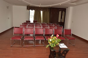 Un salón de conferencias con filas de sillas rojas, una mesa de vidrio con un florero y un ventanal con cortinas.