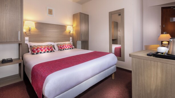 A hotel room with a bed, bedside lamps, a nightstand, a mirror, and a desk with a lamp and a kettle.