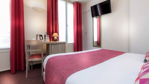 A hotel room with a bed, a desk with a chair, a TV mounted on the wall, and red curtains.