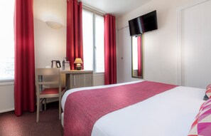 Une chambre d’hôtel avec un lit, un bureau et une chaise, un téléviseur fixé au mur et des rideaux rouges.