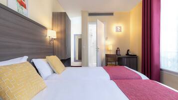 Hotelkamer met twee bedden, een bureau, een stoel, een raam met gordijnen en een deur die leidt naar een andere kamer.