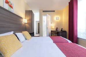 Une chambre d’hôtel avec deux lits, un bureau, une chaise, une fenêtre avec des rideaux et une porte donnant sur une autre pièce.