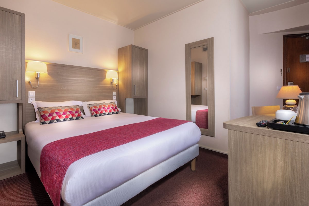 Een hotelkamer met een bed, nachtlampjes, een nachtkastje, een spiegel en een bureau met een lamp en een waterkoker.