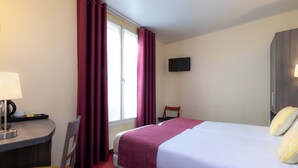 A hotel room with a bed, a desk with a lamp, a chair, a TV on the wall, and a window with curtains.