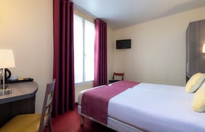 Une chambre d’hôtel avec un lit, un bureau avec une lampe, une chaise, un téléviseur fixé au mur et une fenêtre avec des rideaux.