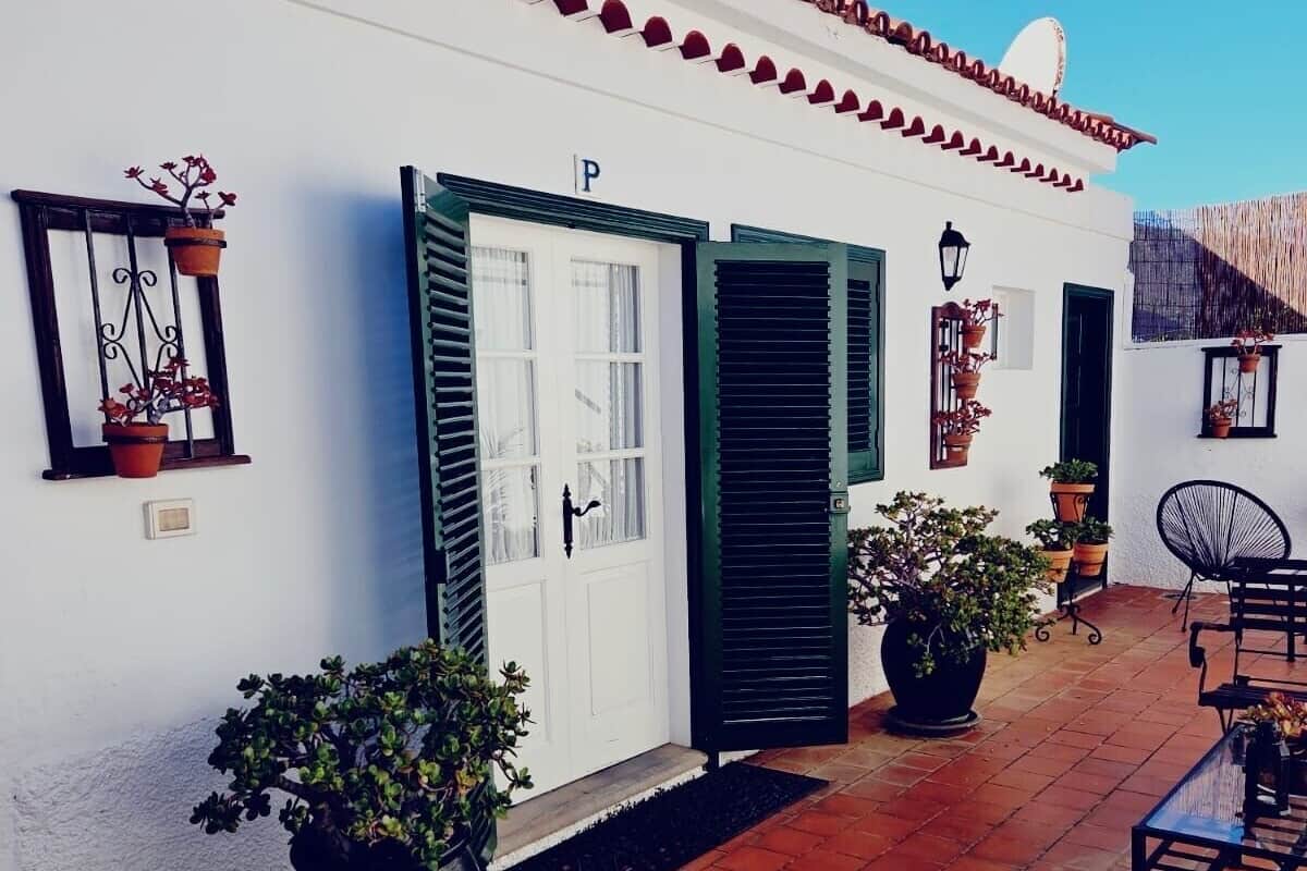 Un edificio blanco con una puerta marcada 'P' y una ventana con una planta. Hay una silla negra y una mesa en el patio.
