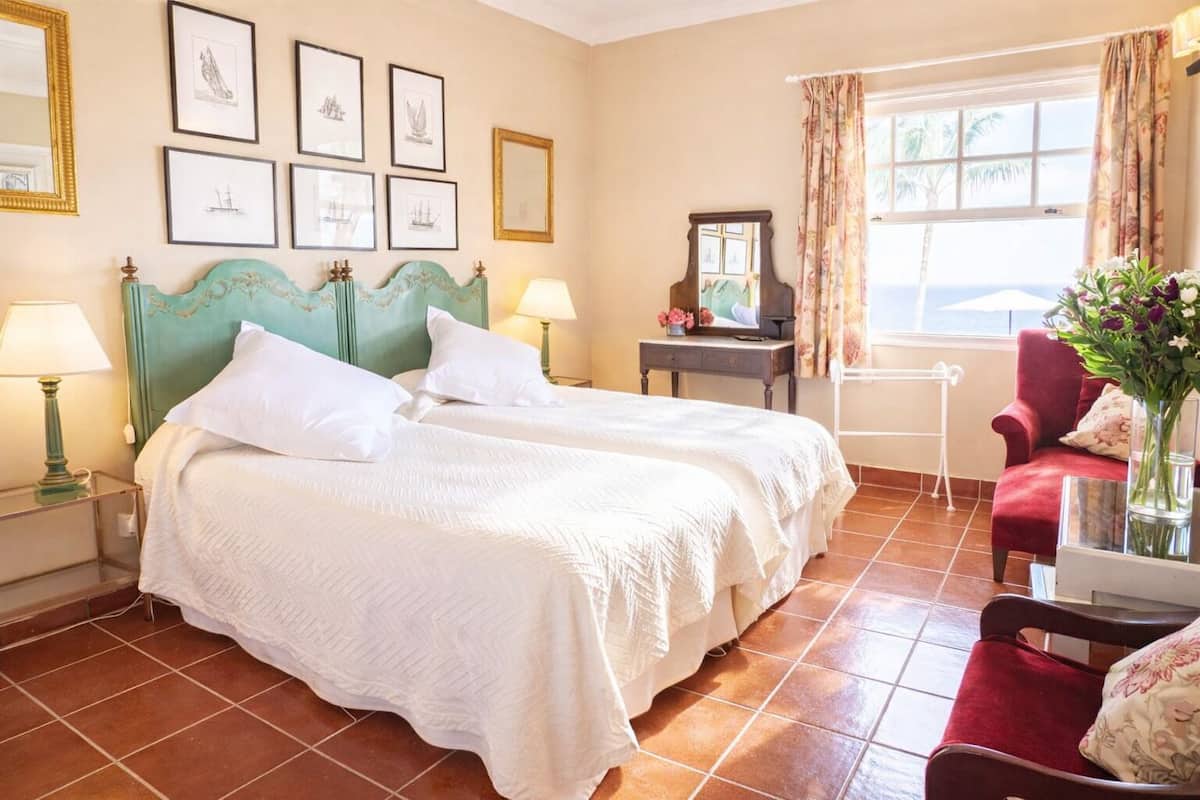 Habitación de hotel con dos camas, un sillón rojo, una mesita con un florero y cuadros en la pared.