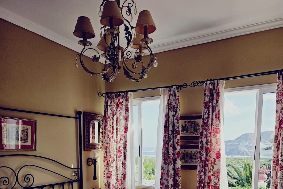 Habitación de hotel con dos camas, una araña, cortinas florales y vistas al paisaje.