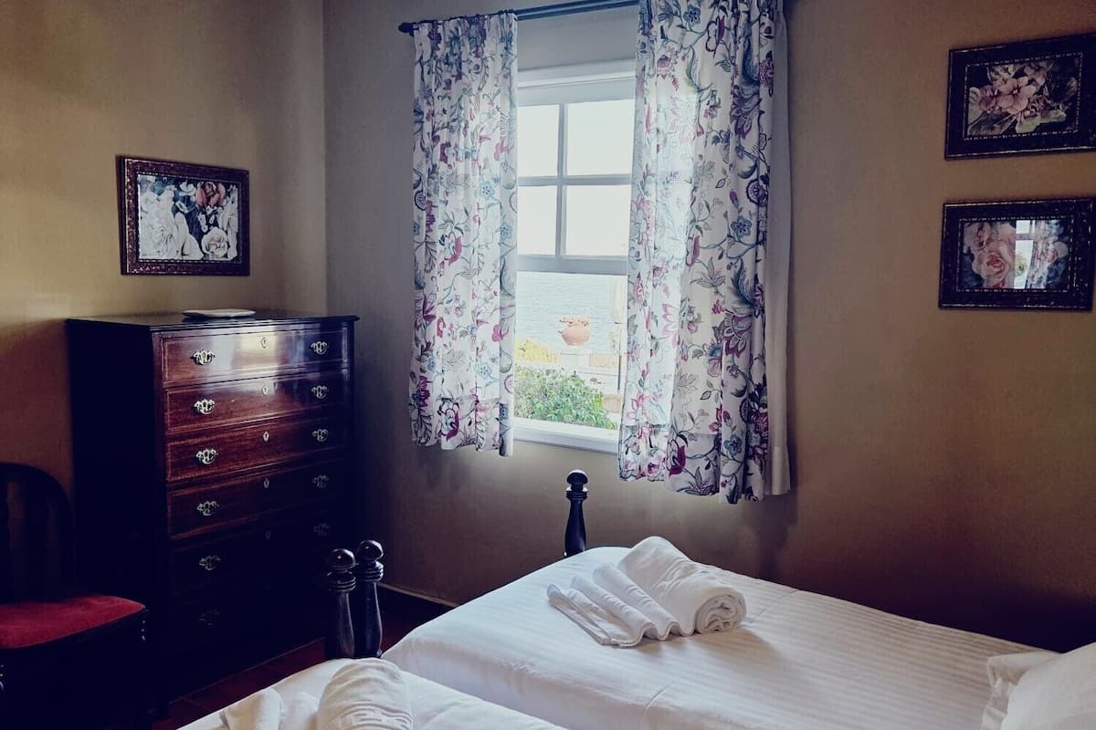 Habitación de hotel con dos camas, un aparador, una silla y una ventana con cortinas estampadas.