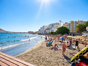 A beach with people swimming and sunbathing, a wooden boardwalk, and buildings in the background. - One bedroom, balcony, Wi-Fi, El Médano centre (El Médano)