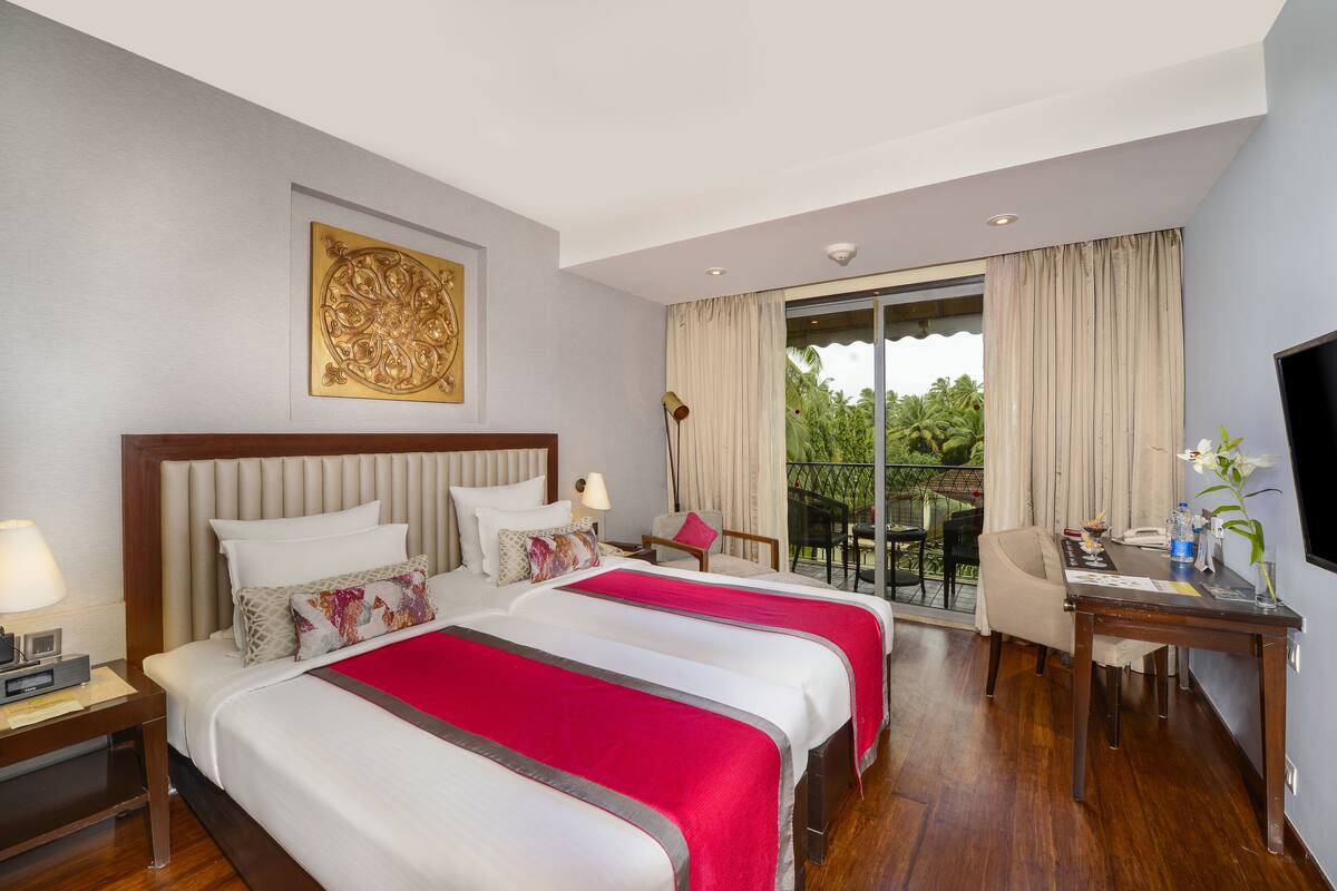 A hotel room with a large bed, a desk, a chair, a TV, and a balcony with a view of greenery.