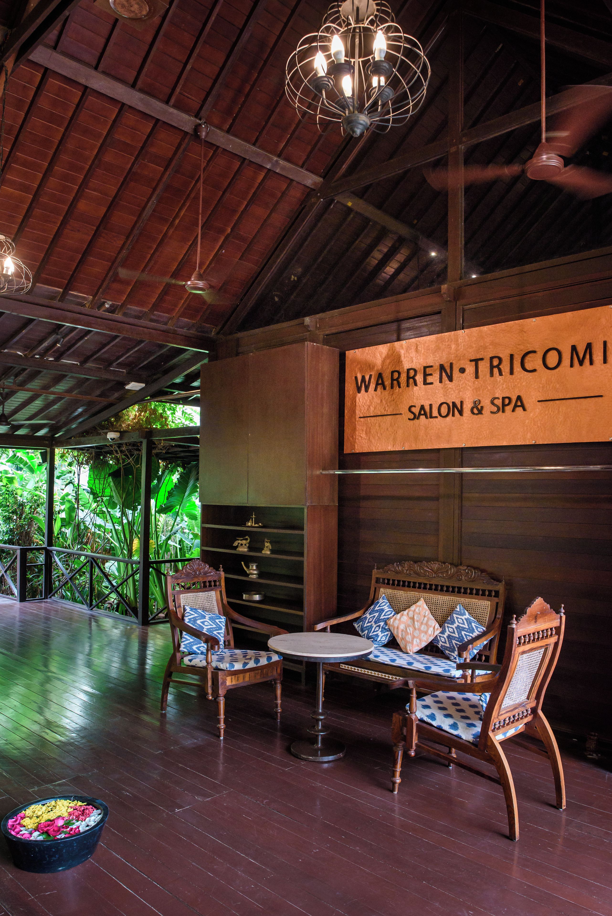 Ett träpanelat tak, en skylt med texten ”Warren Tricomini Salon & Spa”, trästolar med mönstrade kuddar och ett litet bord.
