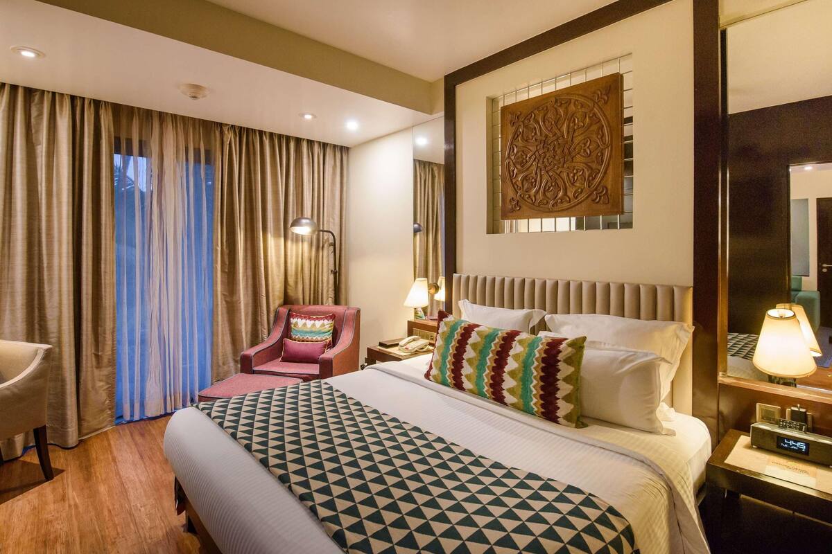 A hotel room with a large bed, a chair, a desk with a lamp, and a wall-mounted artwork.