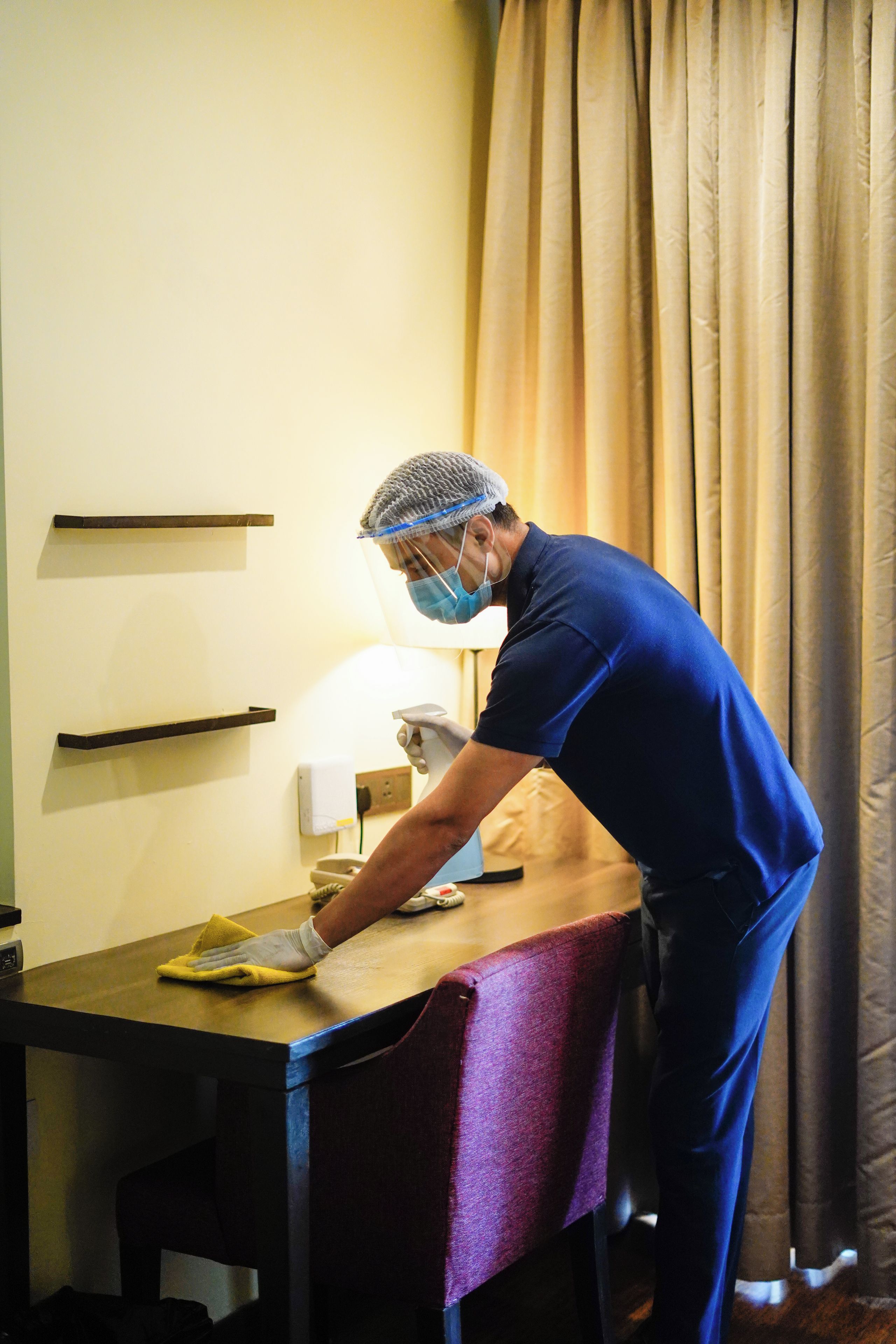 A person cleaning a table in a room with a curtain, a wall-mounted shelf, and a lamp.