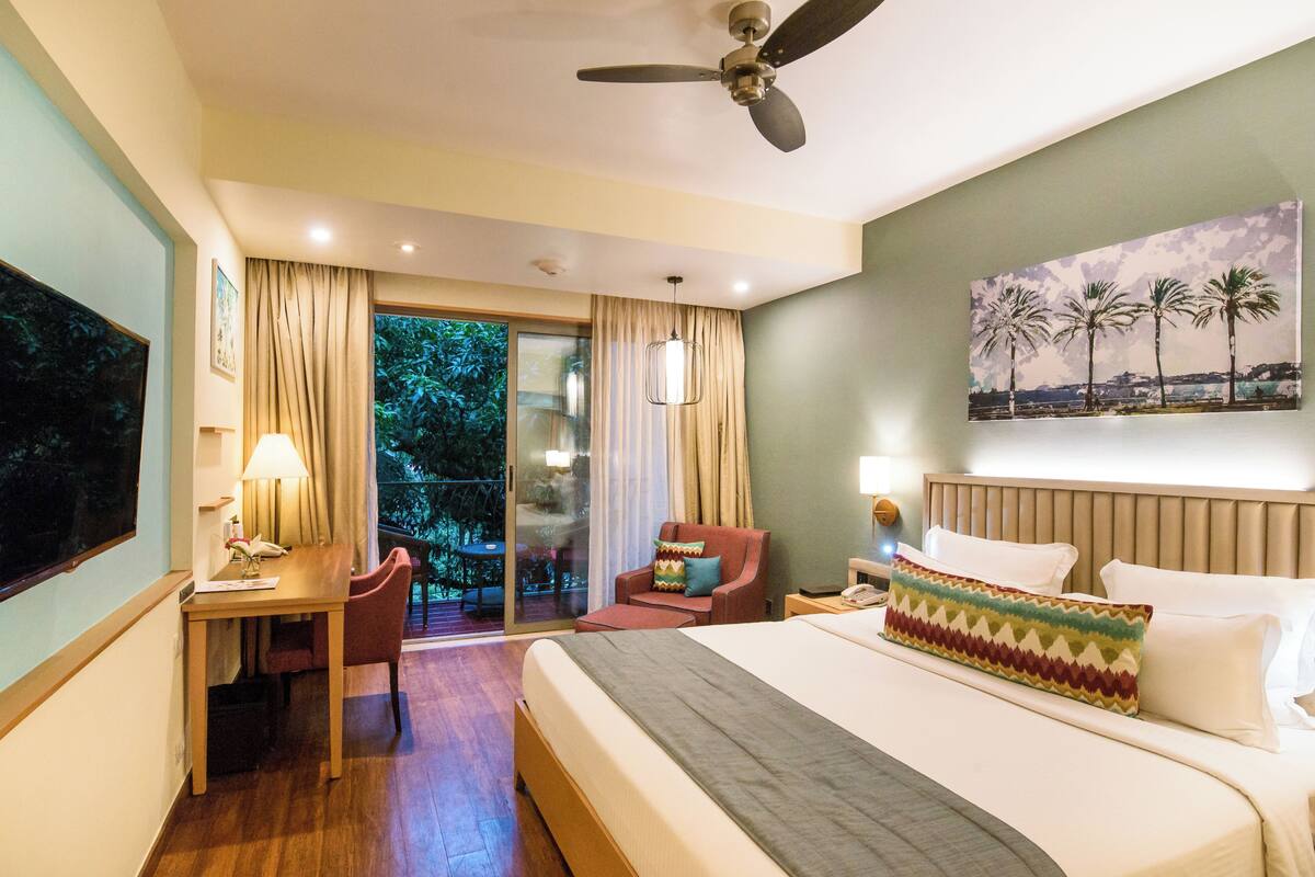 A hotel room with a bed, a desk, a chair, a balcony with a view of trees, and a ceiling fan.