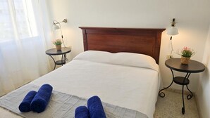Een net opgemaakt bed met witte beddengoed en twee opgerolde blauwe handdoeken, een houten hoofdbord, twee kleine tafels met lampen en potplanten, en een raam met dunne gordijnen.