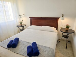 A neatly made bed with white bedding and two rolled blue towels, a wooden headboard, two small tables with lamps and potted plants, and a window with sheer curtains. - 3 bedrooms, swimming pool, parking, 100 metres from the sea, Wi-Fi (Los Cristianos)