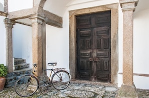A stone doorway with a dark wooden door, a bicycle parked outside, and a potted plant to the left. - Casas De Romaria (Mora)