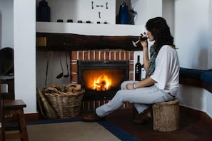 A person sitting by a fireplace with a fire, a basket of wood, and a bottle of wine. - Casas De Romaria (Mora)
