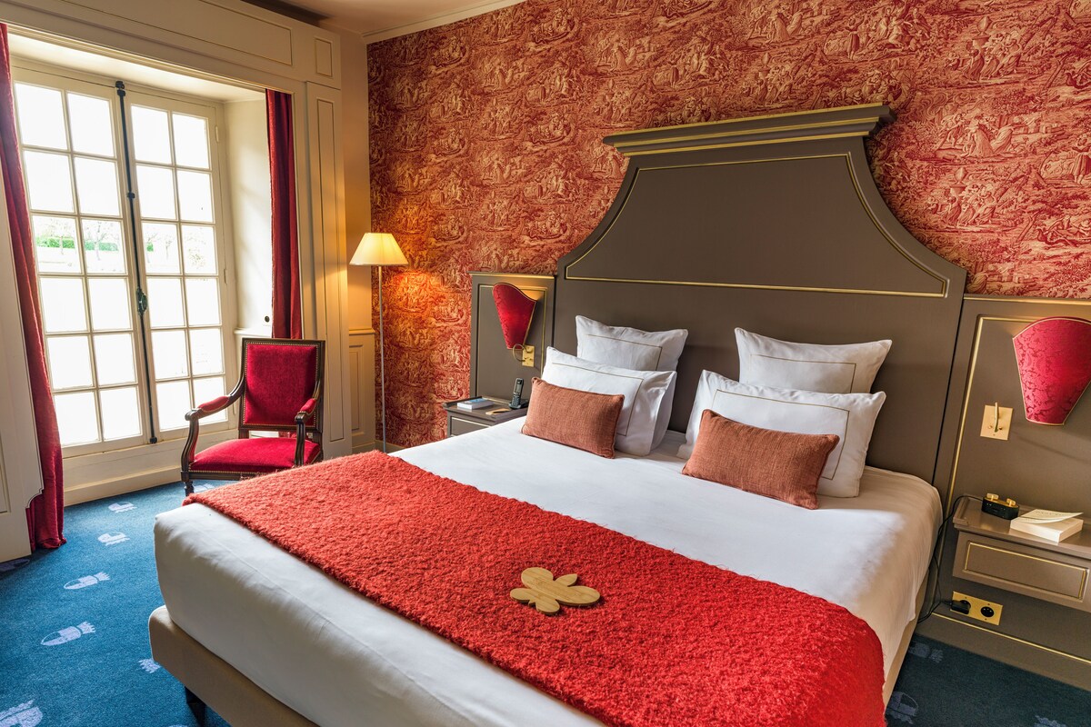 Habitación de hotel con una cama grande, una silla roja y un papel tapiz estampado.