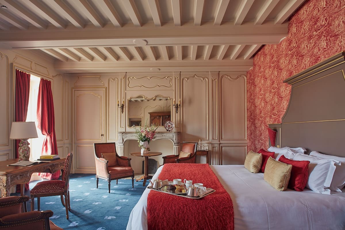 Quarto de hotel com uma cama grande, uma mesa com luminária, duas cadeiras, uma mesinha com um vaso e uma bandeja com itens de café da manhã.
