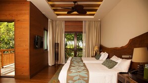 A double bed with a leaf-patterned runner, a wooden headboard, a ceiling fan, and a view of greenery through a large window.