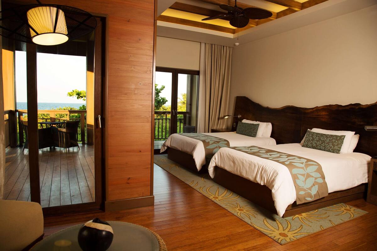 Habitación de hotel con dos camas, suelo de madera, balcón con mobiliario exterior y vista al mar.