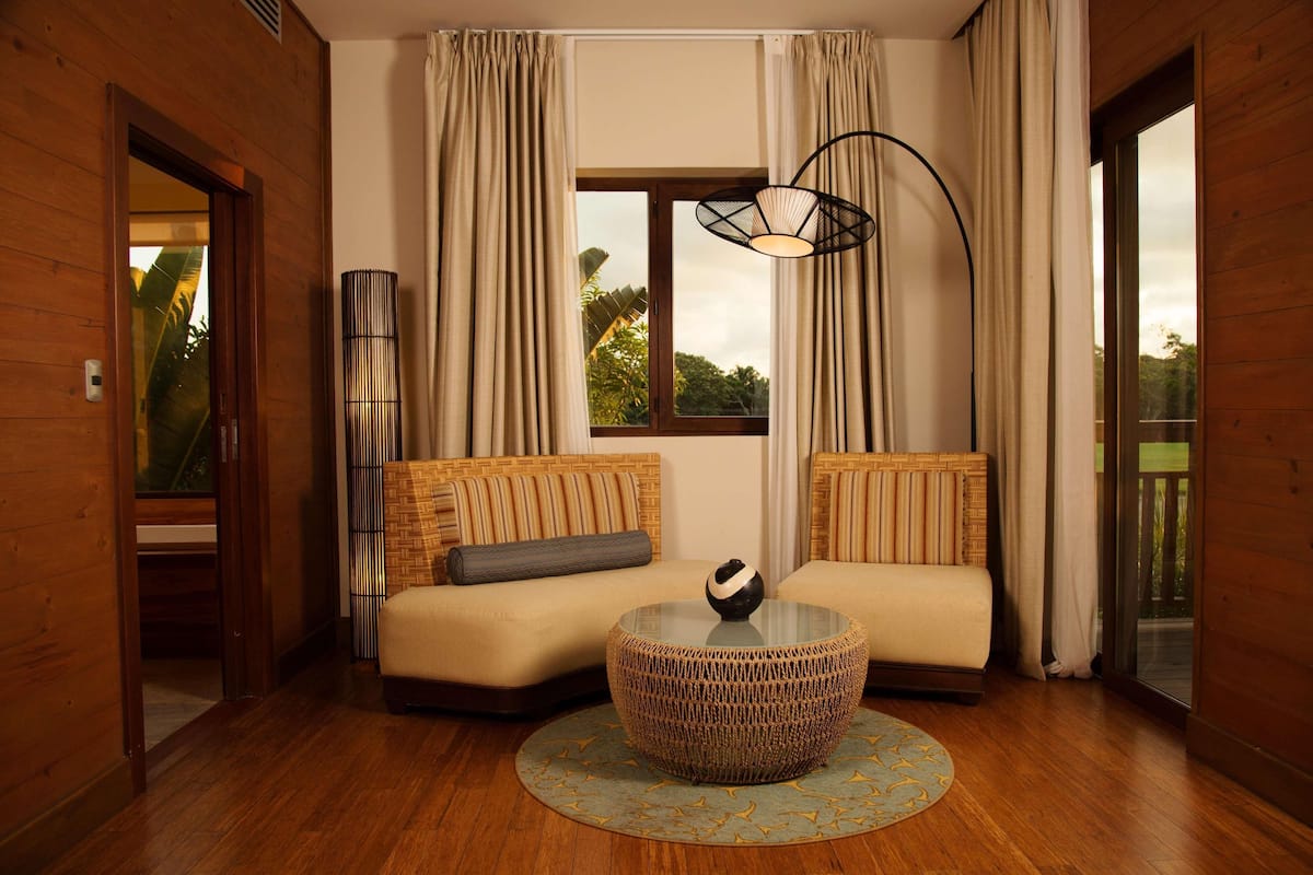 Una habitación acogedora con un sofá, una mesa de centro redonda de mimbre y un amplio ventanal con cortinas.