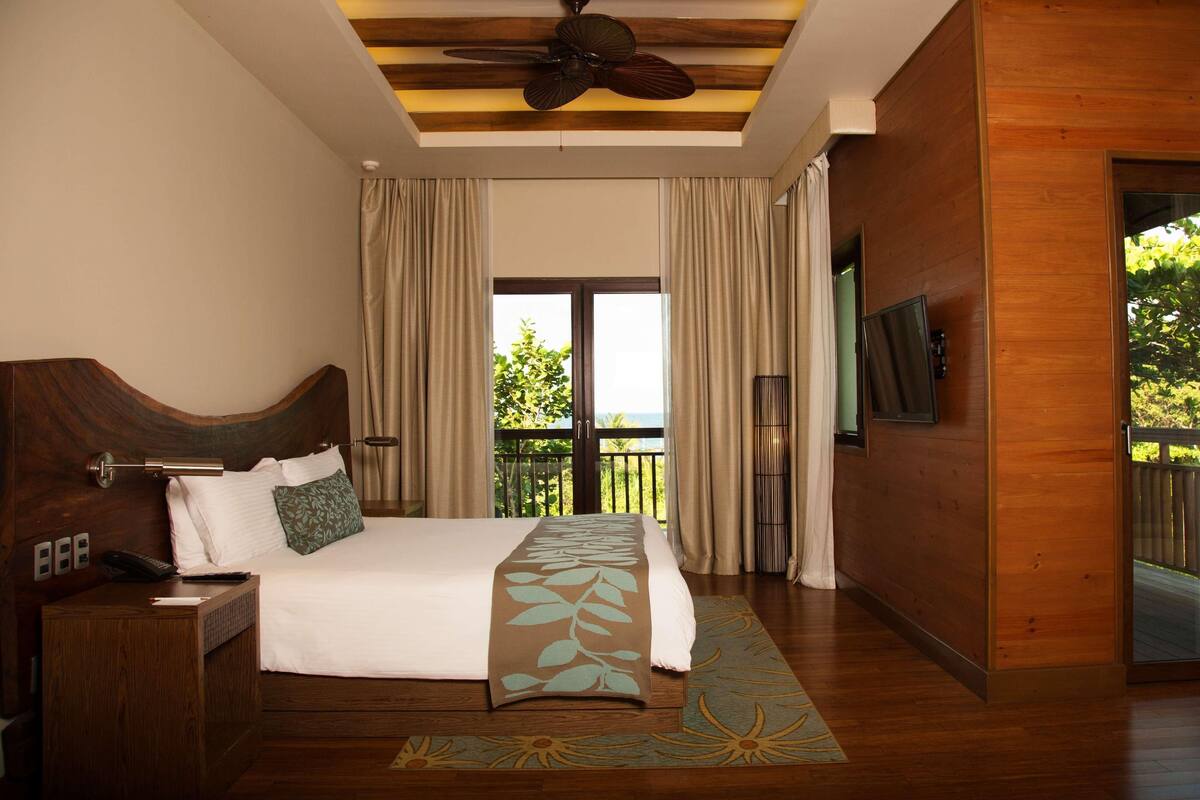 Un dormitorio con cama, cabecera de madera, cortinas, ventilador de techo, televisor y vistas al balcón.