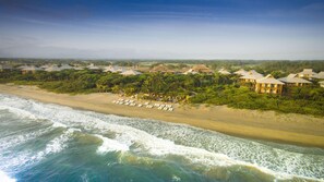 An aerial view of a beach resort with multiple bungalow-style buildings, a sandy beach with lounge chairs, and ocean waves.