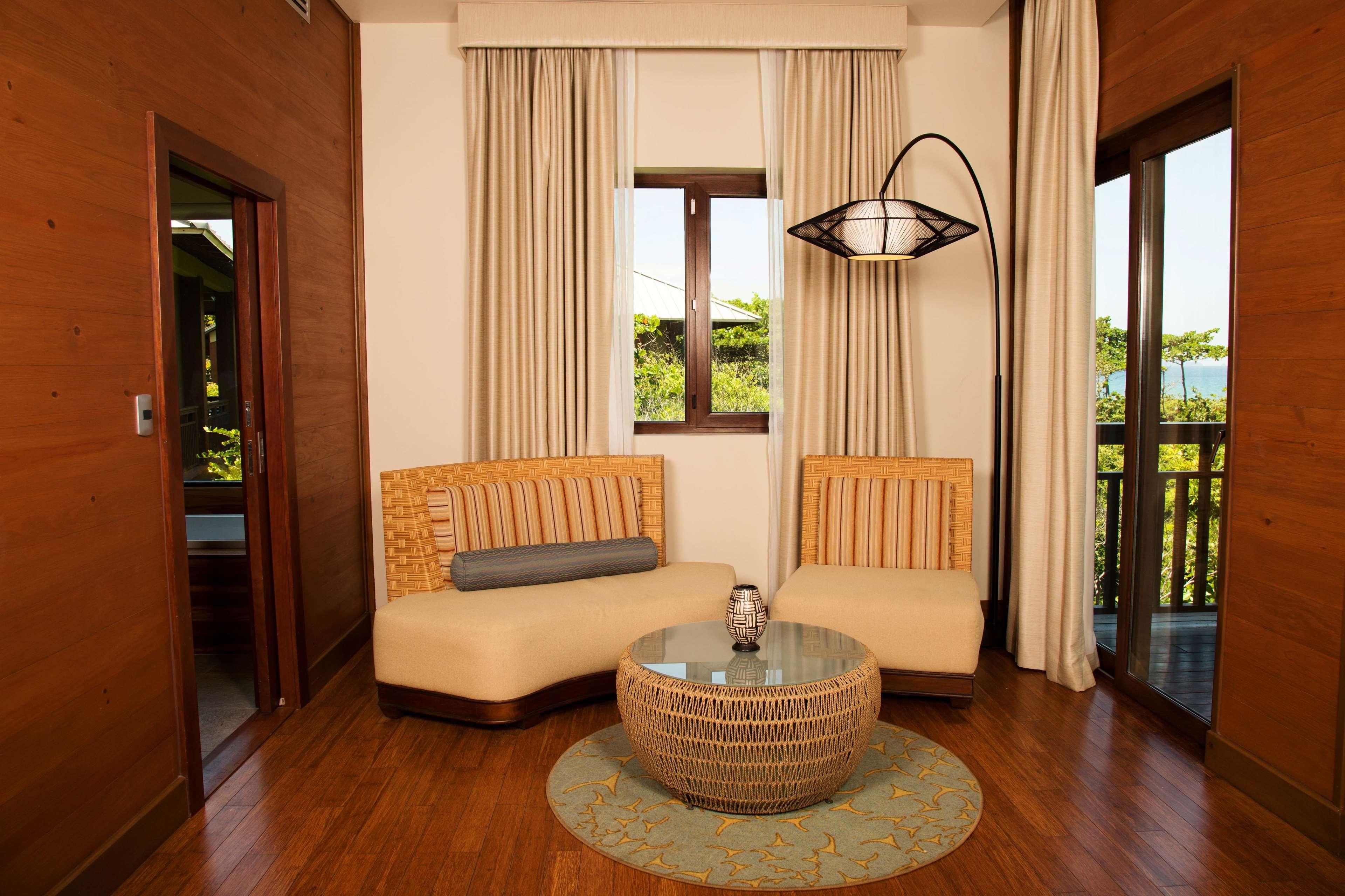 A room with a wooden floor, two sofas with cushions, a round wicker coffee table, a window with curtains, and a view of greenery and a body of water.