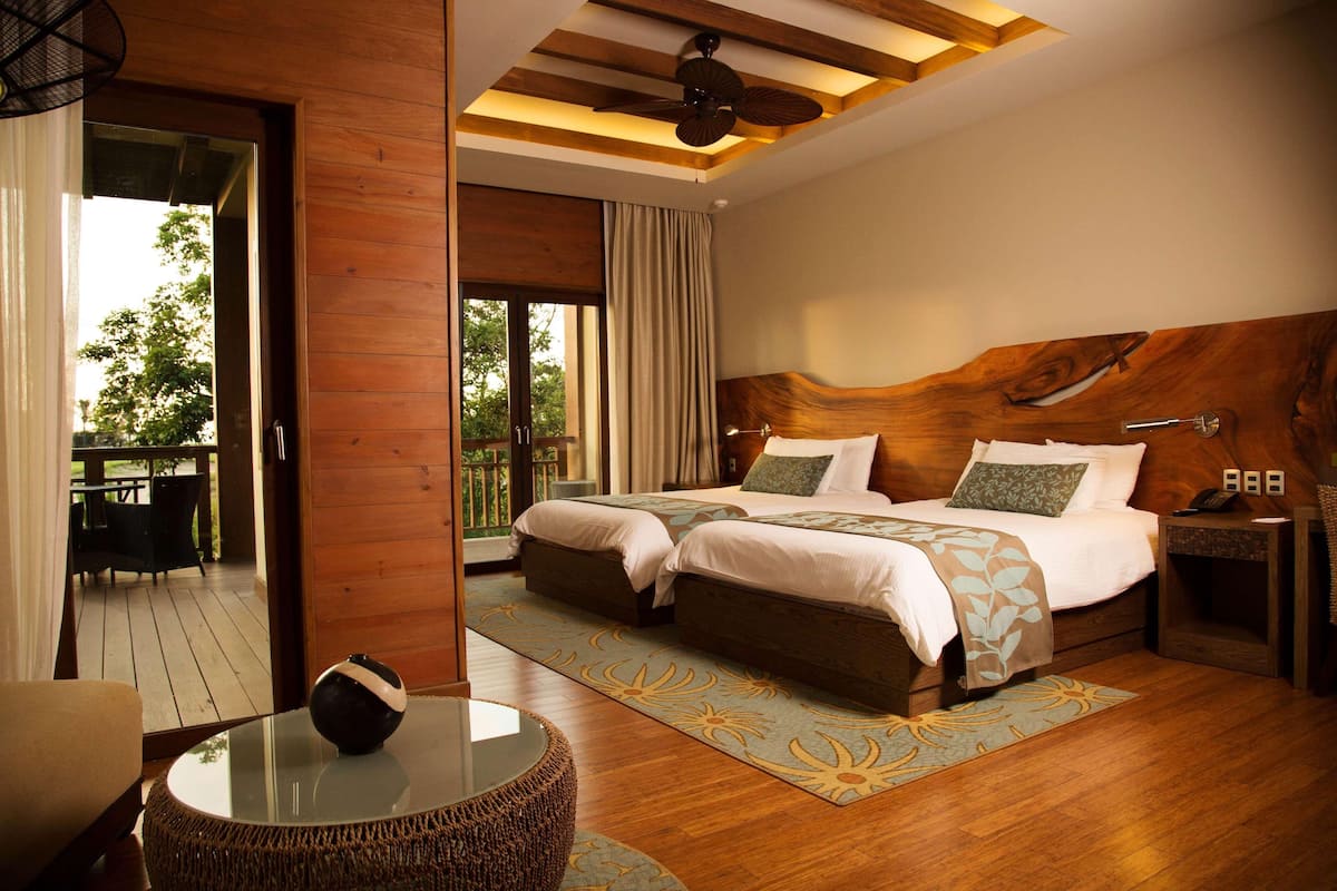 A bedroom with a large bed, a glass table, a wicker chair, and a view of the outdoors.