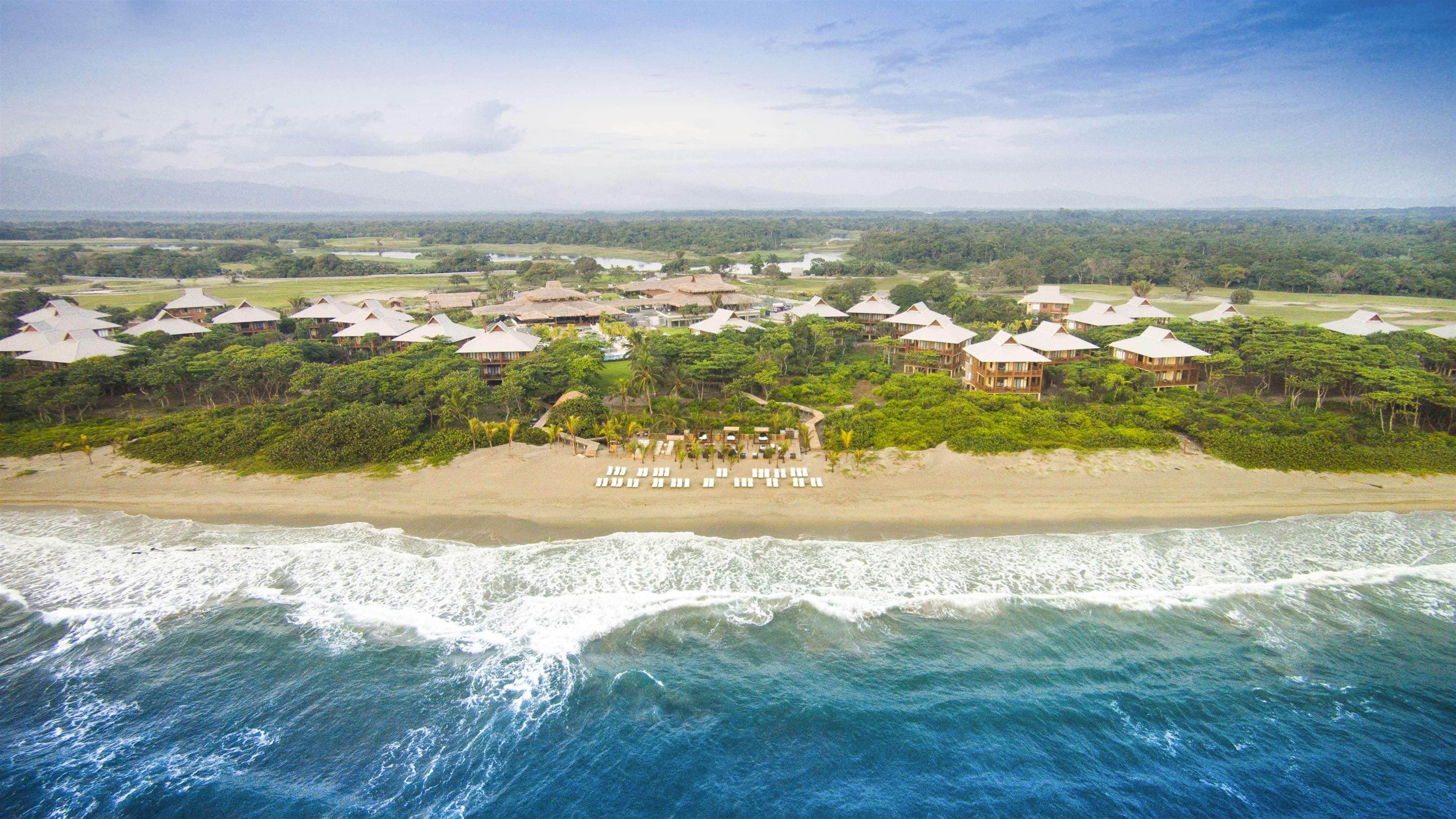 An aerial view of a coastal resort with beachfront villas, a beach with sun loungers, and a backdrop of lush greenery and distant mountains.