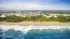 An aerial view of a coastal resort with beachfront villas, a beach with sun loungers, and a backdrop of lush greenery and distant mountains. - Indura Beach & Golf Resort, Curio Collection (Tela)