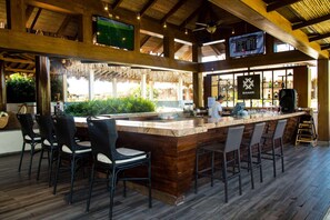 A bar area with a wooden bar counter, black bar stools, and a large screen displaying sports.